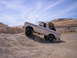 1989 Ford Bronco - A white Ford Bronco navigates a steep gravel incline under a partly cloudy sky.