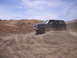 1989 Ford Bronco - A dark blue 1989 Ford Bronco with a tan top navigates a dusty, gravelly incline under a partly cloudy sky.