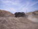 1989 Ford Bronco - A dark pickup truck navigates a dusty, gravelly terrain under a partly cloudy sky.