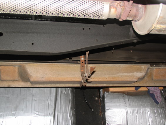 Underside of a vehicle showing a rusty bracket attached to a frame rail, with exhaust pipe above