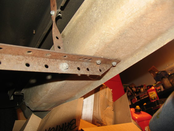 Close-up of a rusted metal bracket with multiple holes, attached to the underside of a vehicle. Cardboard boxes are visible below