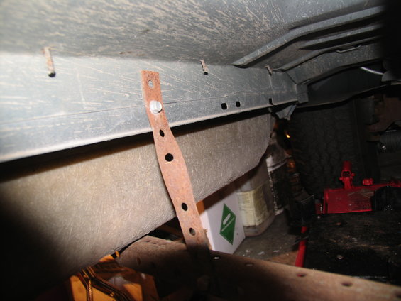 Underbody of a vehicle showing a rusty metal bracket attached to the frame, with a tire and red jack visible in the background