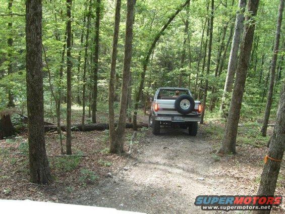1992 Ford Bronco with spare tire on the back driving on a dirt road through trees