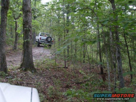 White SUV driving up a dirt path in a wooded area. Spare tire mounted on the back