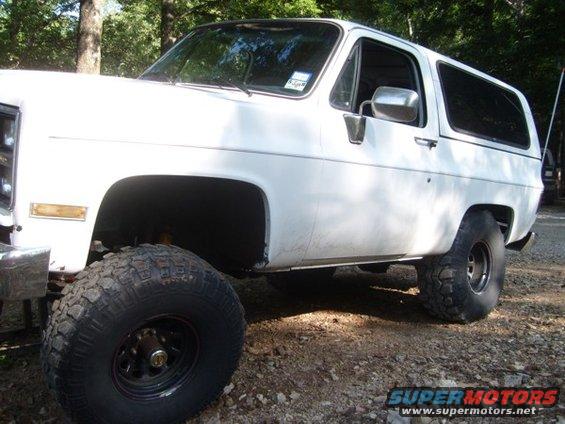 White SUV with large off-road tires, lifted suspension, parked on gravel. Side view shows two doors and tinted rear windows