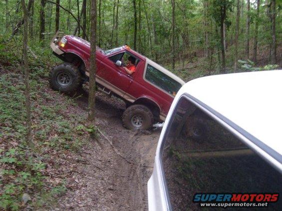 Red SUV with lifted suspension is going up a steep, muddy trail in the woods. Another white vehicle is in the foreground