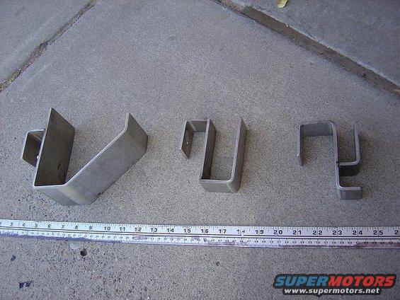 Three metal brackets of varying sizes laid out on a concrete surface next to a ruler