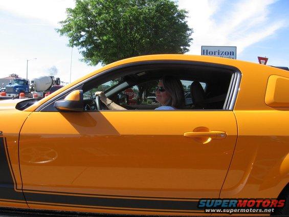 Side view of an orange coupe with black racing stripes and a woman driving