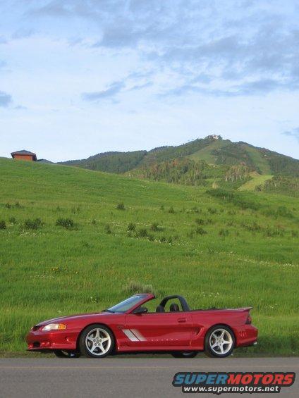 Red convertible Mustang parked on the side of a road with a grassy hill and mountains in the background