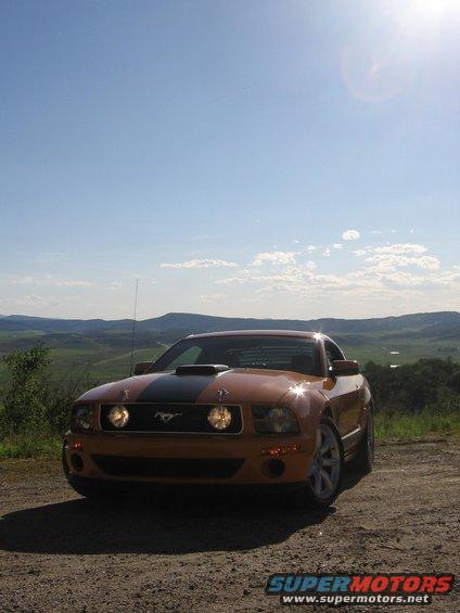 Orange Ford Mustang with black hood stripe and scoop, parked on a dirt road with rolling hills in background