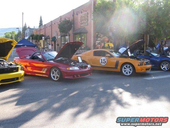 Yellow, red, orange, and blue Mustangs lined up with hoods open. Orange one has a '15' on the door