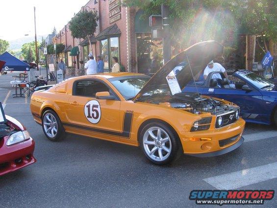 Orange Ford Mustang with racing stripes and a large number 15 on the door, hood open, parked on a street