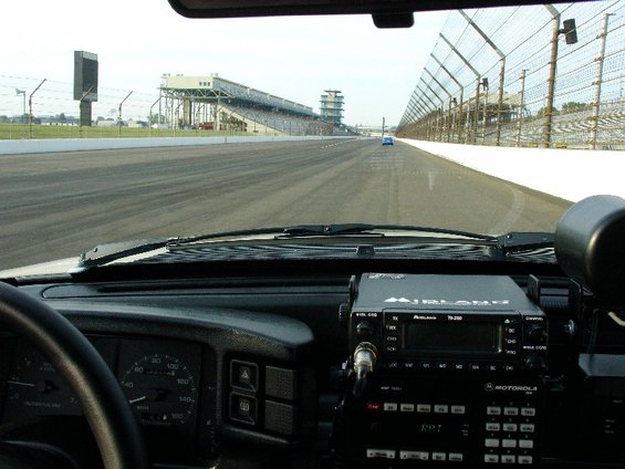 View from inside a vehicle looking out at the Indianapolis Motor Speedway track with grandstands and a distant blue car