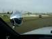 1993 Ford Mustang - A red car drives on a racetrack, seen from the perspective of another vehicle's interior, with a large chrome side mirror in the foreground.