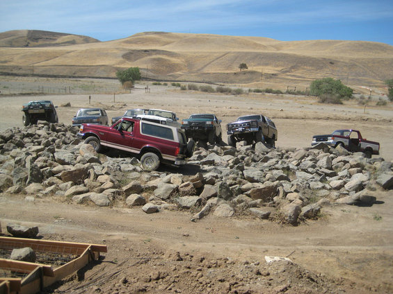 Several off-road trucks and SUVs are positioned on and around a large pile of rocks, with dry, hilly terrain in the background