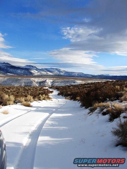 Snowy road winding through a desolate landscape with snow-capped mountains under a blue sky with clouds