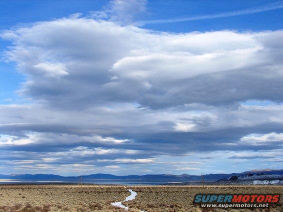 Cloudy sky over a dry, brushy landscape with a winding stream and distant mountains