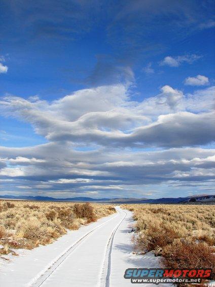 snowy road in a dry, brushy landscape under a cloudy blue sky