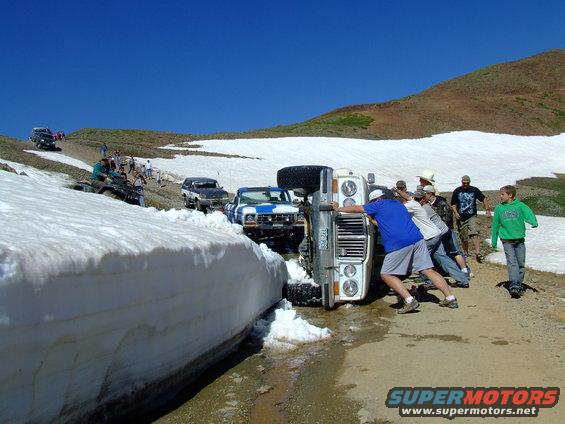 Overturned 1987 Ford Bronco on snowy road, people pushing it upright