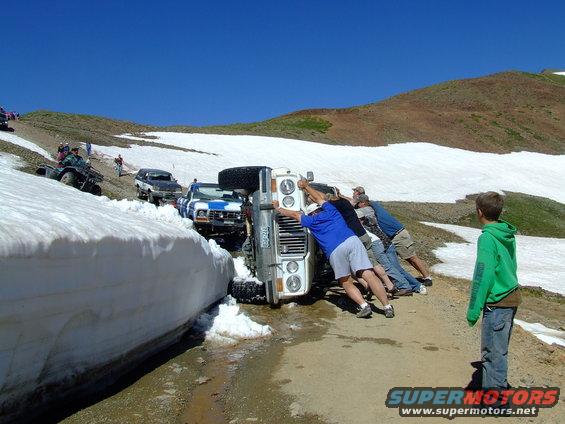 1987 Ford Bronco overturned on a mountain road, people pushing it upright. Another SUV and ATV visible