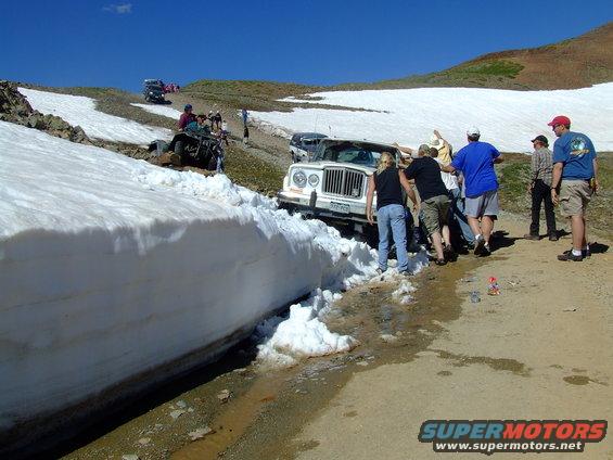 White Jeep stuck in a snowbank on a dirt road, people pushing to free it. Other vehicles visible further up the road