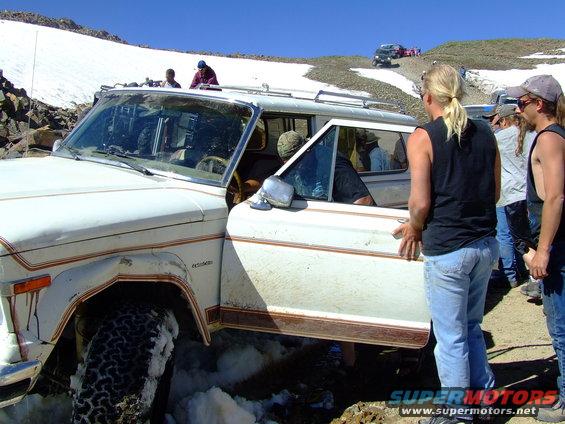 White SUV stuck in snow, door open, people around. Appears to be a Jeep Grand Wagoneer