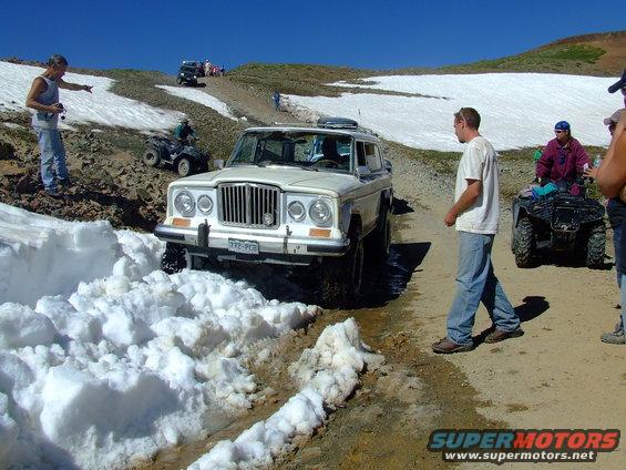 White Jeep Wagoneer with a prominent grille stuck in snow on a dirt road, people and ATVs nearby