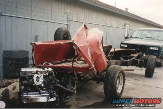 Wrecked red pickup truck chassis with engine and transmission removed, spare tire on top. Another dark SUV is parked behind it