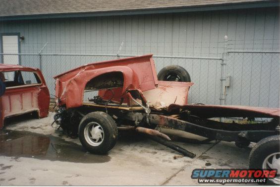 Damaged red pickup truck chassis with a separate cab in the background. The truck is missing its body panels and engine