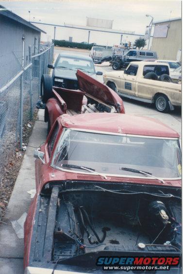 Red pickup truck with hood off, parked next to a dark pickup. Another yellow pickup is in the background