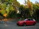 2007 Mazda 3 - A red 2007 Mazda 3 hatchback is parked on the side of a winding road with trees in the background.
