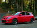 2007 Mazda 3 - A red 2007 Mazda 3 hatchback is parked on a gravel lot with trees and a road in the background.