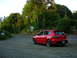 2007 Mazda 3 - A red 2007 Mazda 3 hatchback is parked on a gravel shoulder next to a tree-lined road.