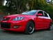 2007 Mazda 3 - A red 2007 Mazda 3 hatchback is shown from a three-quarter front angle on a gravel surface with trees in the background.
