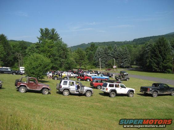 Group of SUVs and trucks parked on a grassy hill with trees and mountains in the background