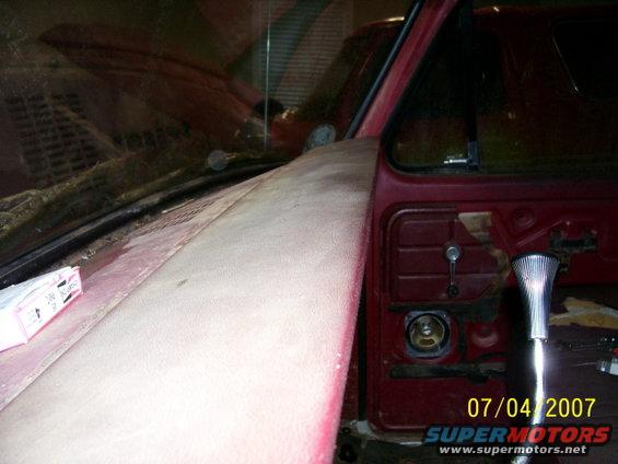 Red vehicle interior with door panel removed, showing a chrome door handle and speaker grille. The hood is visible, covered in dust. Another red vehicle is reflected in the windshield