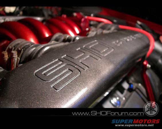 Dark gray engine intake manifold with raised "SHO" emblem and red hoses