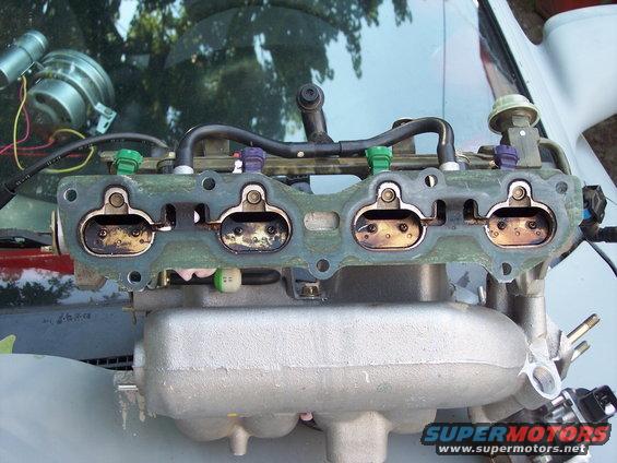 Close-up of a car's intake manifold with fuel injectors attached. The manifold has four openings with butterfly valves