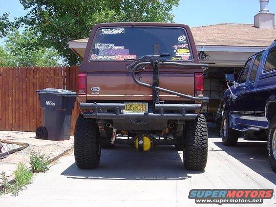 Rear view of a brown 1986 Ford Bronco II with aftermarket bumper and spare tire carrier. Another blue vehicle is parked next to it