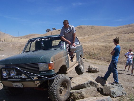 Guy leaning on a green Ford Bronco, tires on rocks, dry hills behind