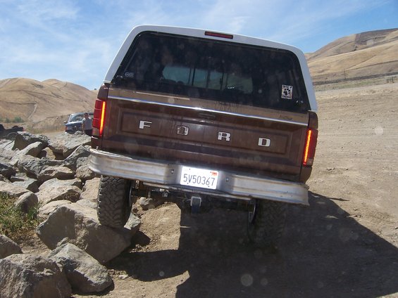 Brown Ford Bronco rear view, white camper shell, off-roading over rocks