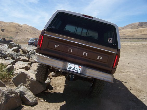 Brown 1987 Ford Bronco with white camper shell driving over rocks. California license plate 5V50367