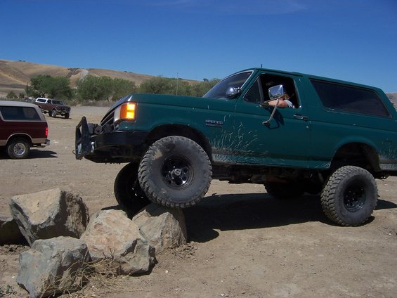 Green 1991 Ford Bronco driving over rocks, with another Bronco and a pickup truck in the background