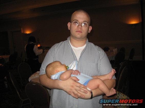 Man in a striped shirt holds a baby wearing a blue onesie and a pacifier. A watermark reads SUPERMOTORS.NET