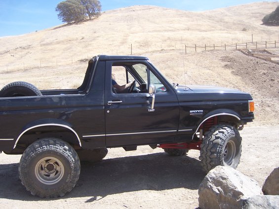 Black Ford Bronco parked on dirt road, spare tire in back. Hills in background