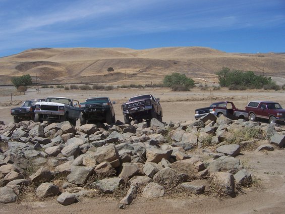 Several trucks are driving over large rocks in a dry, hilly landscape under a blue sky