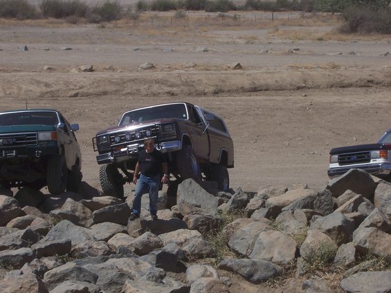 Three lifted trucks navigate a rocky, off-road trail. A man stands near the center truck