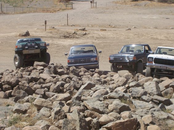 Four Ford trucks and SUVs on a rocky terrain, front view. One green Bronco is lifted with off-road tires