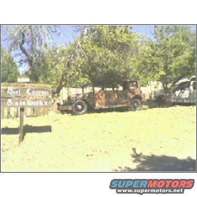 Rusty old car parked outside next to a sign that says "Duel Canyon Bass Works". Another vehicle is partially visible behind it