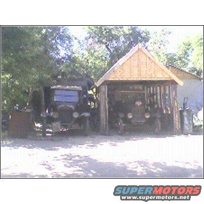 Two dark-colored vintage trucks parked outdoors, one under a wooden carport